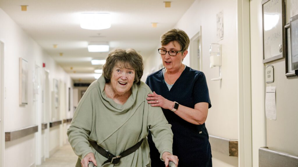A smiling woman walks down the hallway with the assistance of a nurse at a skilled nursing facility